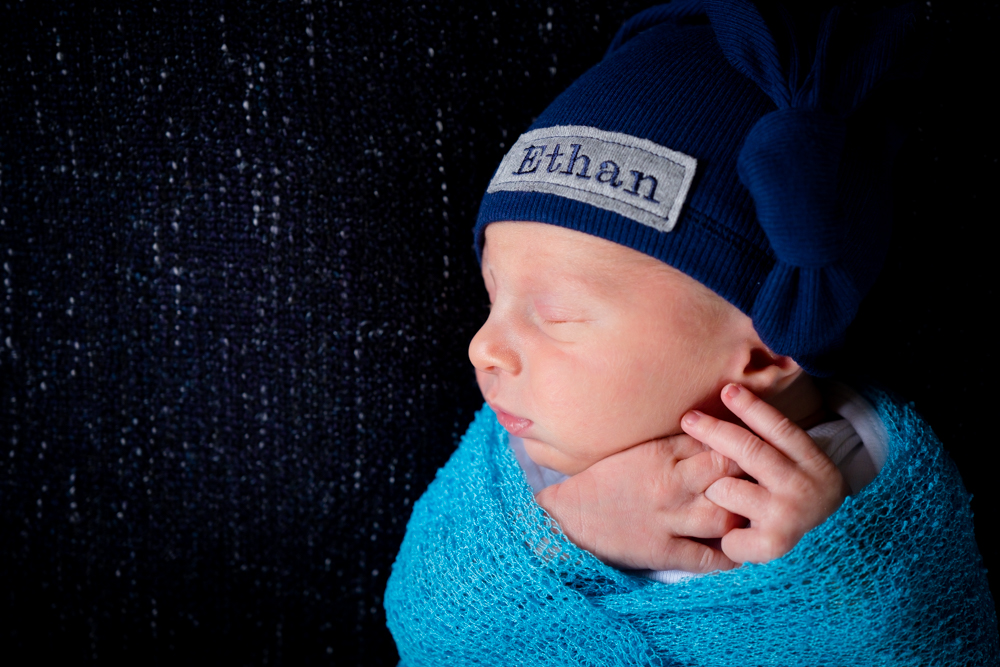 Newborn boy wrapped in a blue wrap with a hat that says "Ethan"
